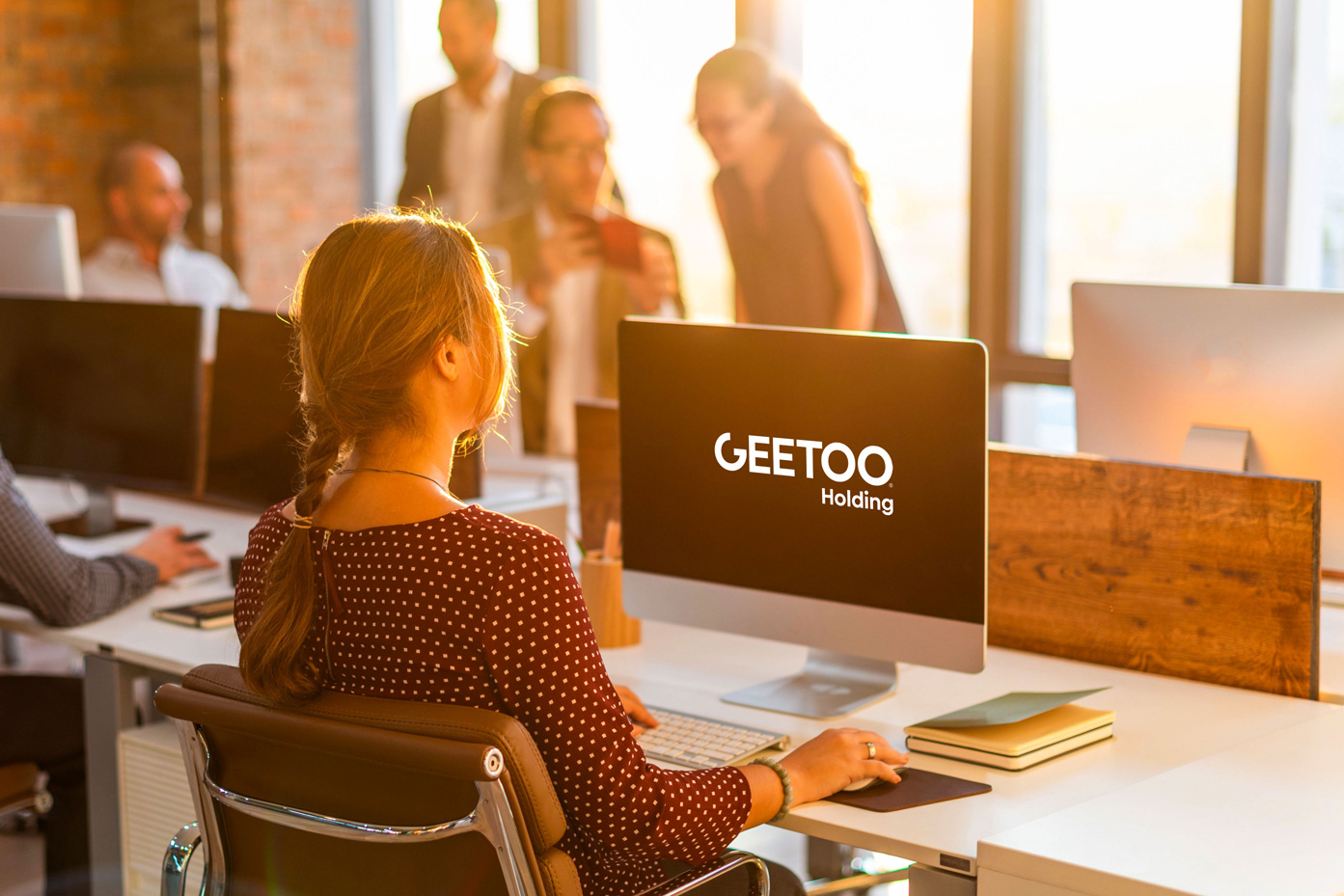 Person sits at a desk in a bright office, facing a computer with 'GEETOO Holding' on screen amid coworkers in the background.