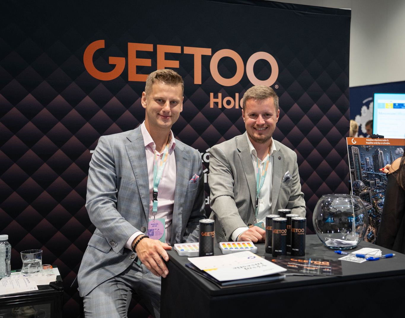 Two men in gray suits sit at a booth with a GEETOO backdrop, smiling at the camera amid brochures, canisters, and a glass bowl on the table.